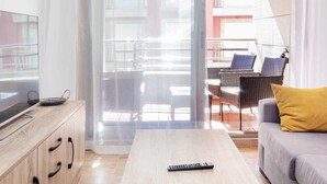 Un salon moderne avec une table basse en bois, un canapé et des fauteuils jaunes. Un téléviseur est posé sur un meuble en bois, et on aperçoit un balcon avec du mobilier d’extérieur.