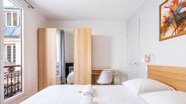 Quarto de hotel com cama, escrivaninha, cadeira, janela e um quadro na parede.
