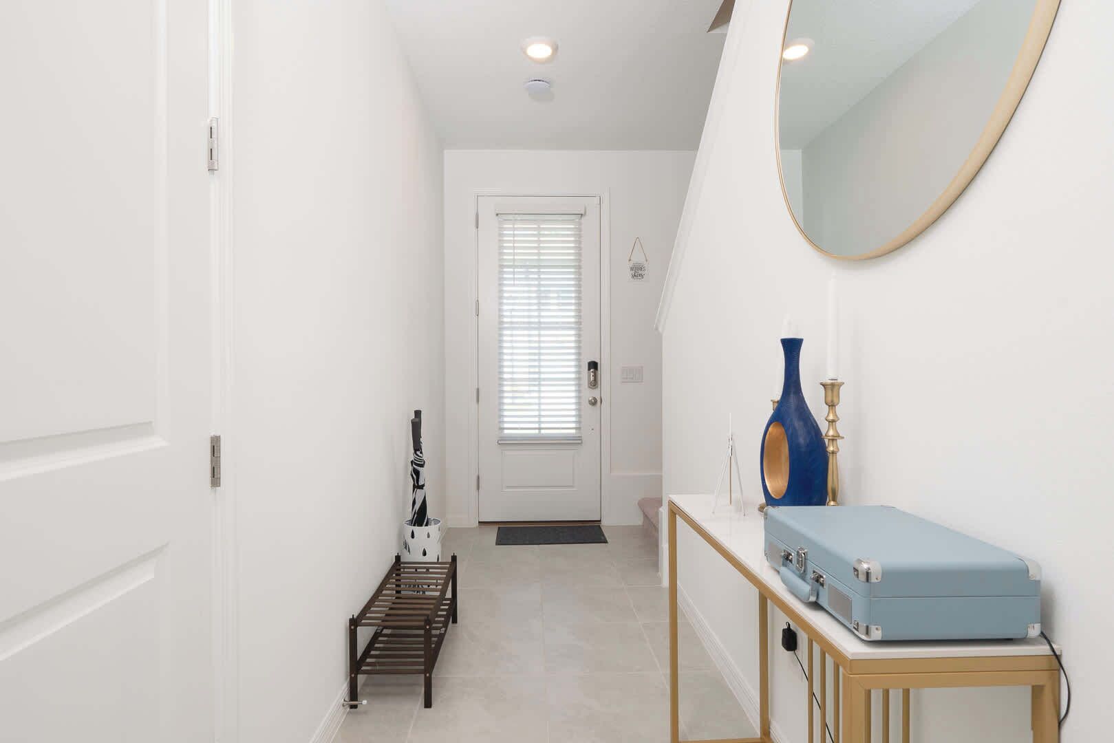 Un couloir avec une porte blanche, un banc, un vase bleu et un miroir au mur.