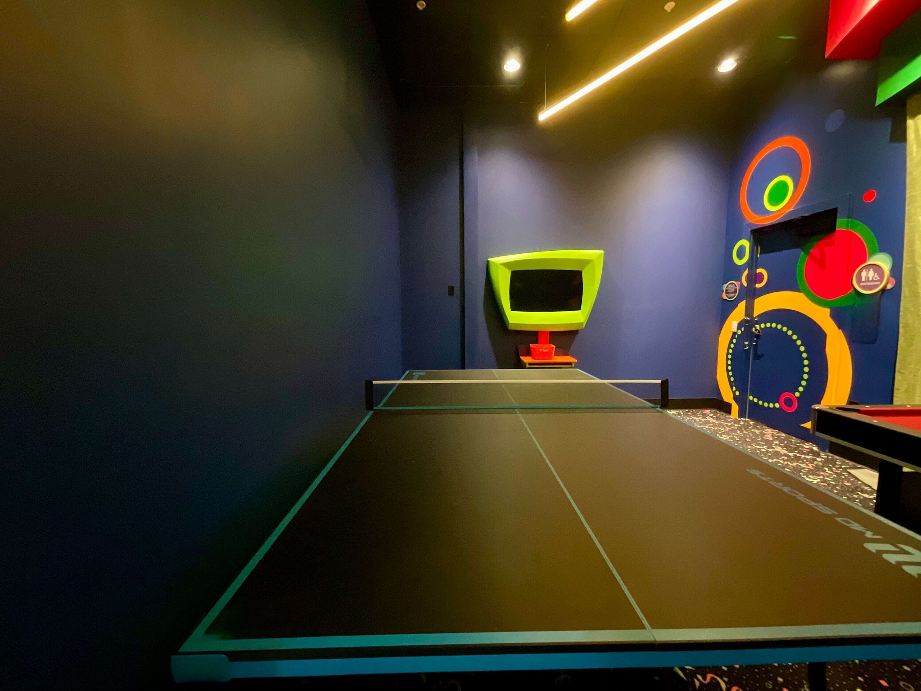 A ping pong table with a neon TV and colorful wall art in a room with dark walls and a modern design.