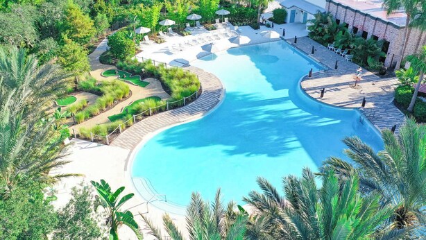 Uma piscina grande e sinuosa cercada por palmeiras e espreguiçadeiras, com um edifício ao fundo.