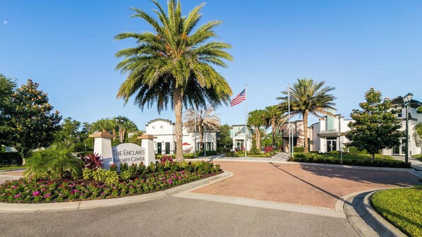 A gated entrance with a sign reading 'The Enclaves Amenity Center,' palm trees, and a clear blue sky. - Brand New 3 BDR Resort Home Packed w/ Amenities! (Davenport)