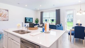 Uma cozinha moderna com uma ilha central, uma área de refeições com cadeiras azuis e uma sala de estar com sofá e televisão.