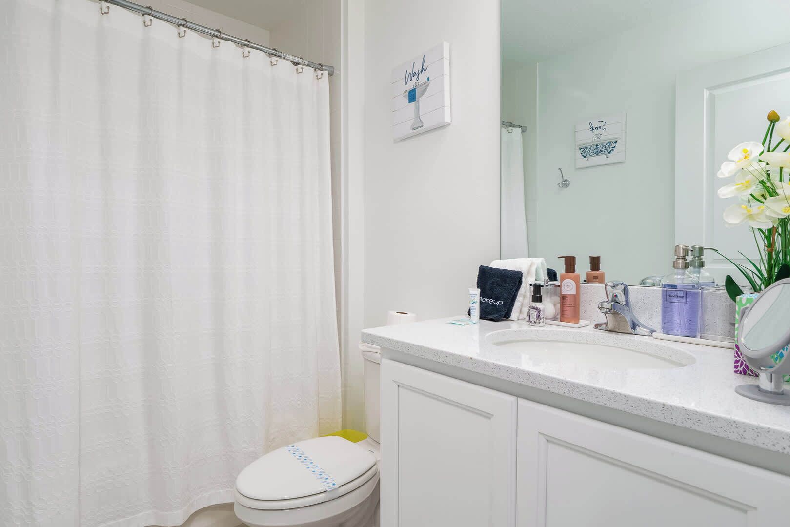 Une salle de bain avec des toilettes blanches, un rideau de douche, un lavabo avec des articles de toilette et un miroir.