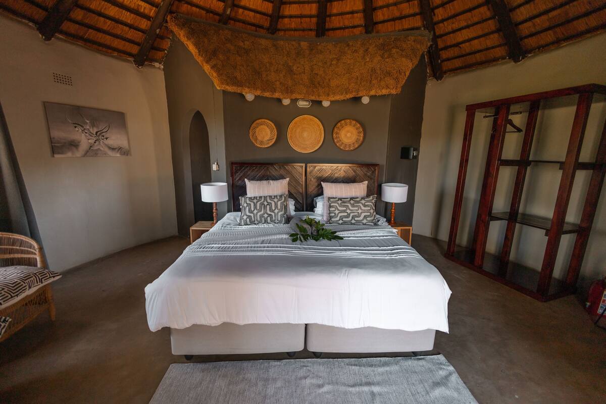 A spacious bedroom with a large bed, two bedside tables, a wooden wardrobe, and a ceiling with a thatched roof.
