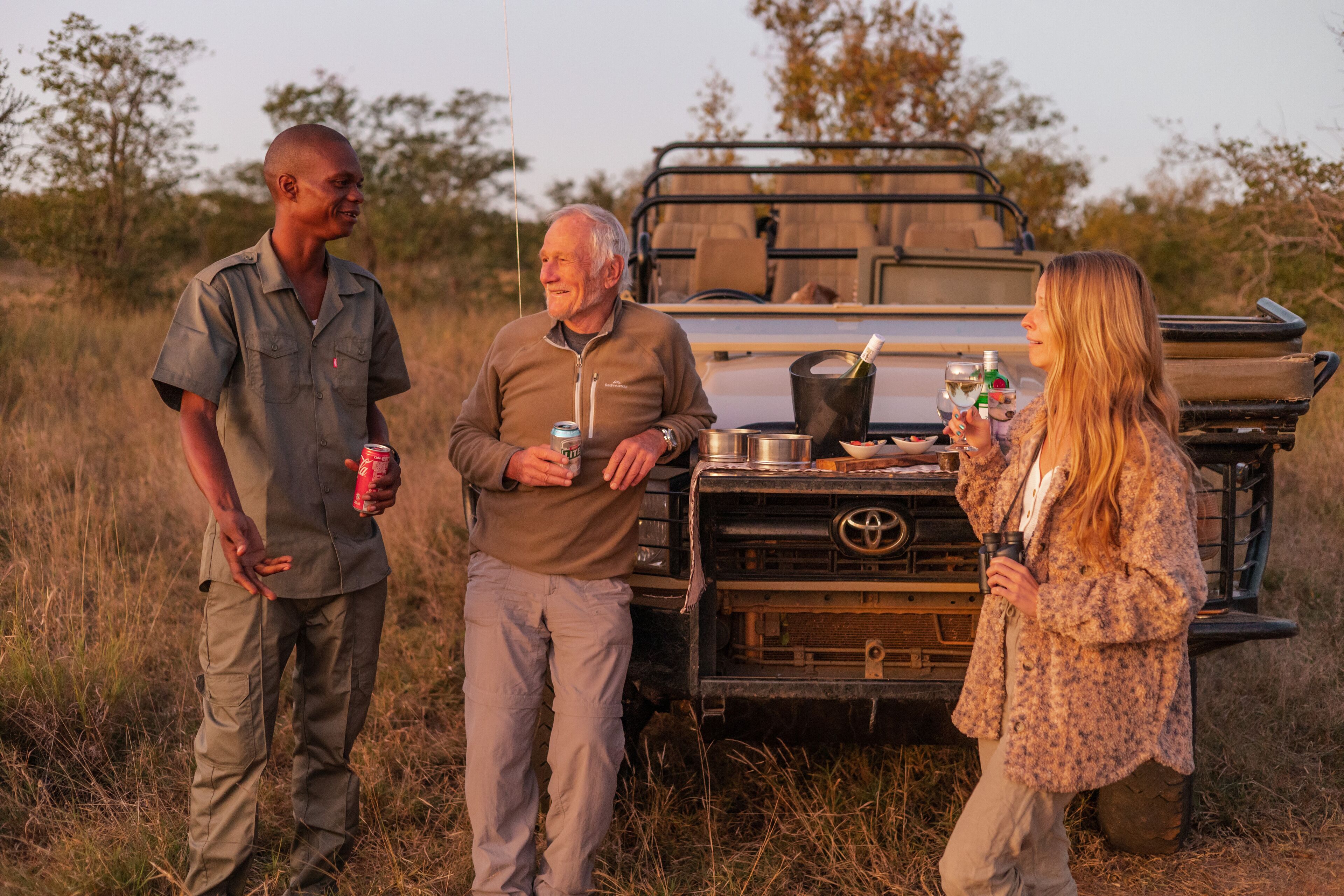 Three people standing in front of a vehicle with a table and drinks.