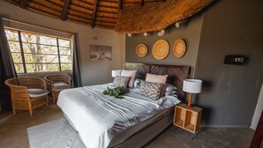 A bedroom with a large bed, two wicker chairs, a wooden nightstand, and a window with a view of trees. - Nsala Wilderness Camp (Hoedspruit)