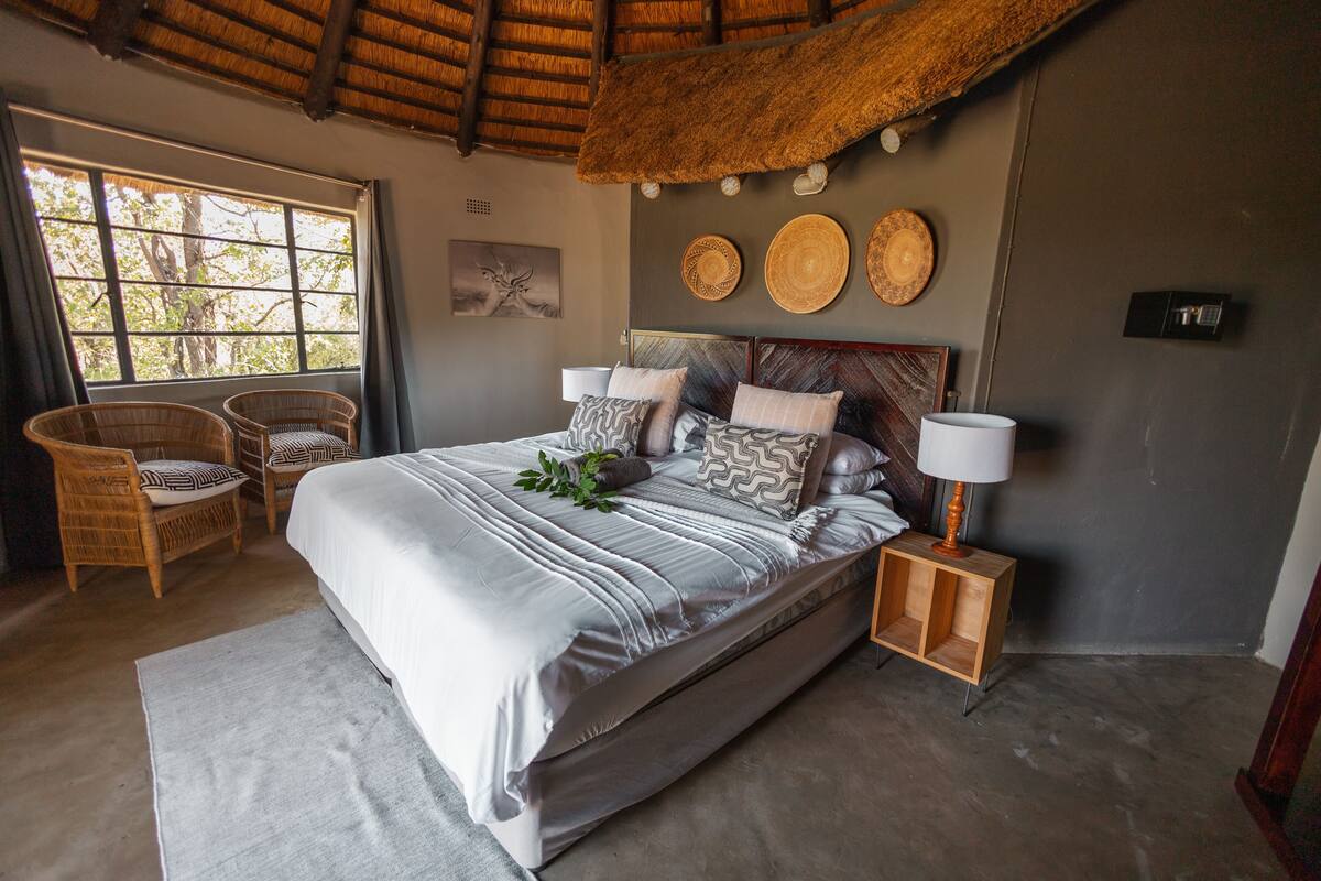 A bedroom with a large bed, two wicker chairs, a wooden nightstand, and a window with a view of trees.
