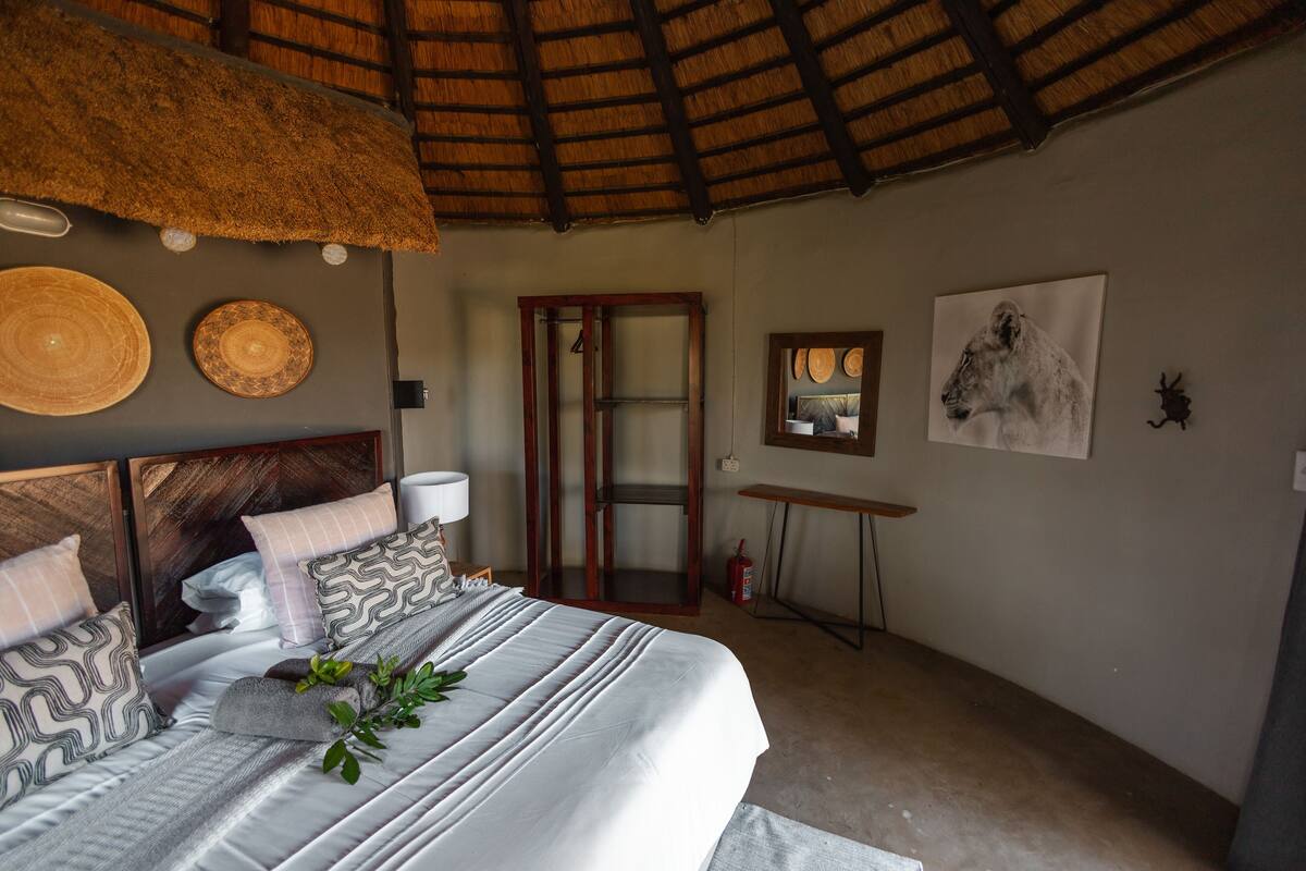 A bedroom with a bed, a thatched roof, a wooden door, and wall decorations including a lion painting.