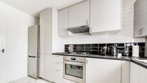 A modern kitchen with white cabinets, a stainless steel refrigerator, a built-in oven, and a black countertop with various appliances. - GuestReady - Urban Comfort near Wembley Stadium (London)