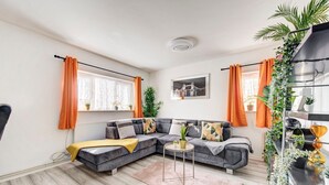 A modern living room with a grey sectional sofa, a small round table, and a pink rug. There are two windows with orange curtains, and a plant shelf on the right. - GuestReady - Urban Comfort near Wembley Stadium (London)