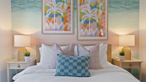 Un lit bien fait, avec du linge de lit blanc, un coussin bleu au point de crochet et deux tableaux abstraits encadrés accrochés au-dessus.