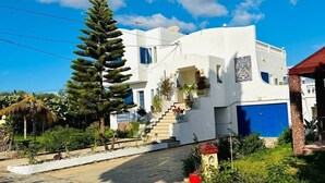 Un edificio blanco con una puerta azul, rodeado de follaje y un camino empedrado.