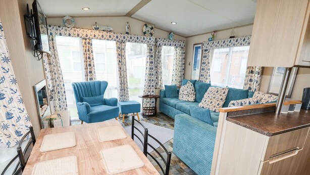 Un intérieur chaleureux avec une table à manger en bois, des sièges recouverts d’un tissu bleu et des rideaux aux motifs marins.