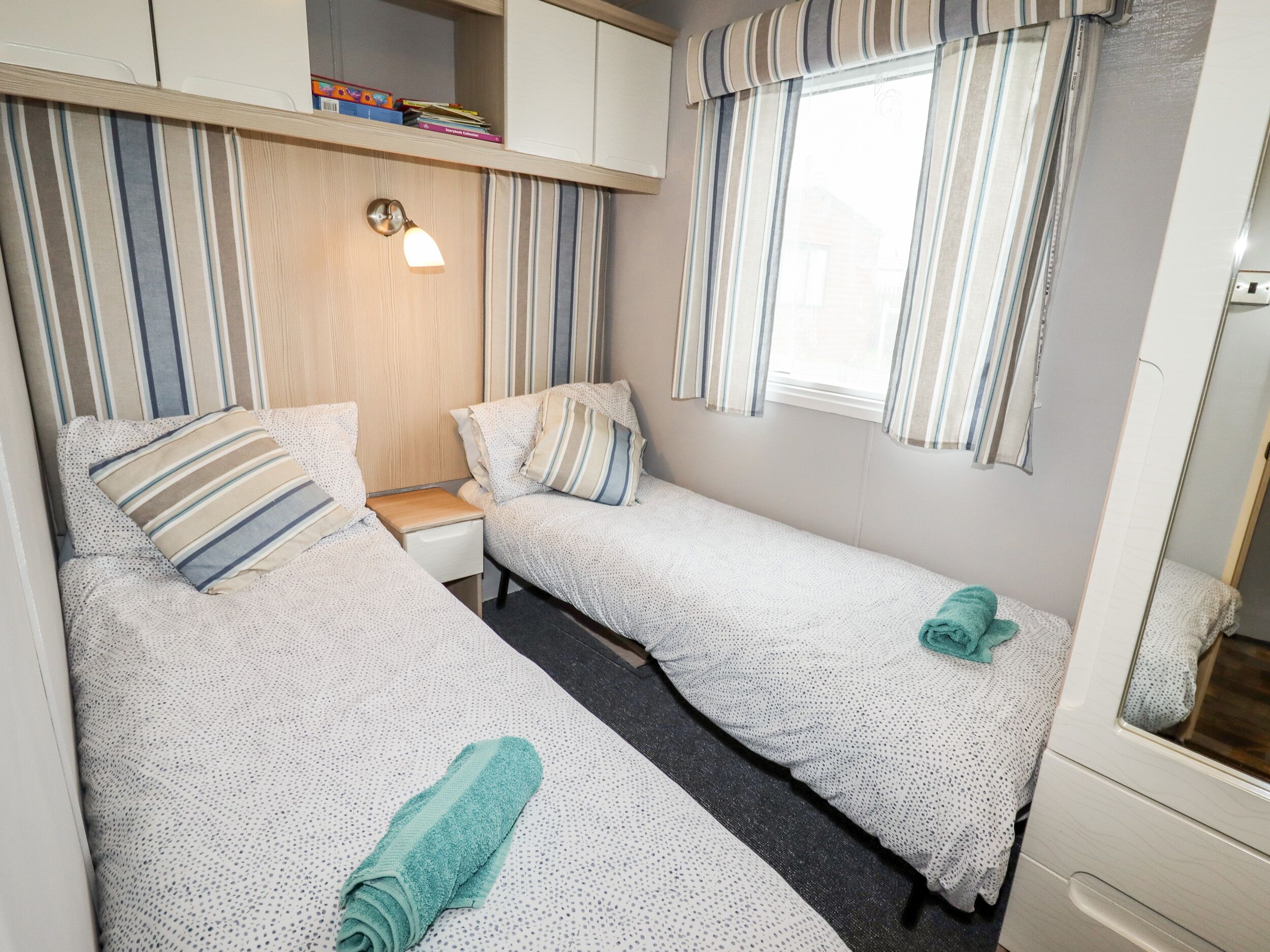 Ein gemütliches Schlafzimmer mit zwei Einzelbetten, einem Nachttisch, einer Lampe, einem Regal mit Büchern und einem Fenster mit gestreifter Gardine.