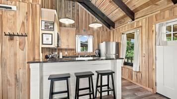 A wooden interior with a kitchen area featuring a bar stool seating, a refrigerator, and a window with curtains.