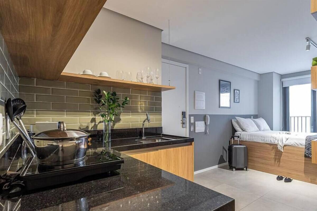 A modern kitchen with a black countertop, a stove, and a plant. There is a bed with a suitcase and a window with a view.
