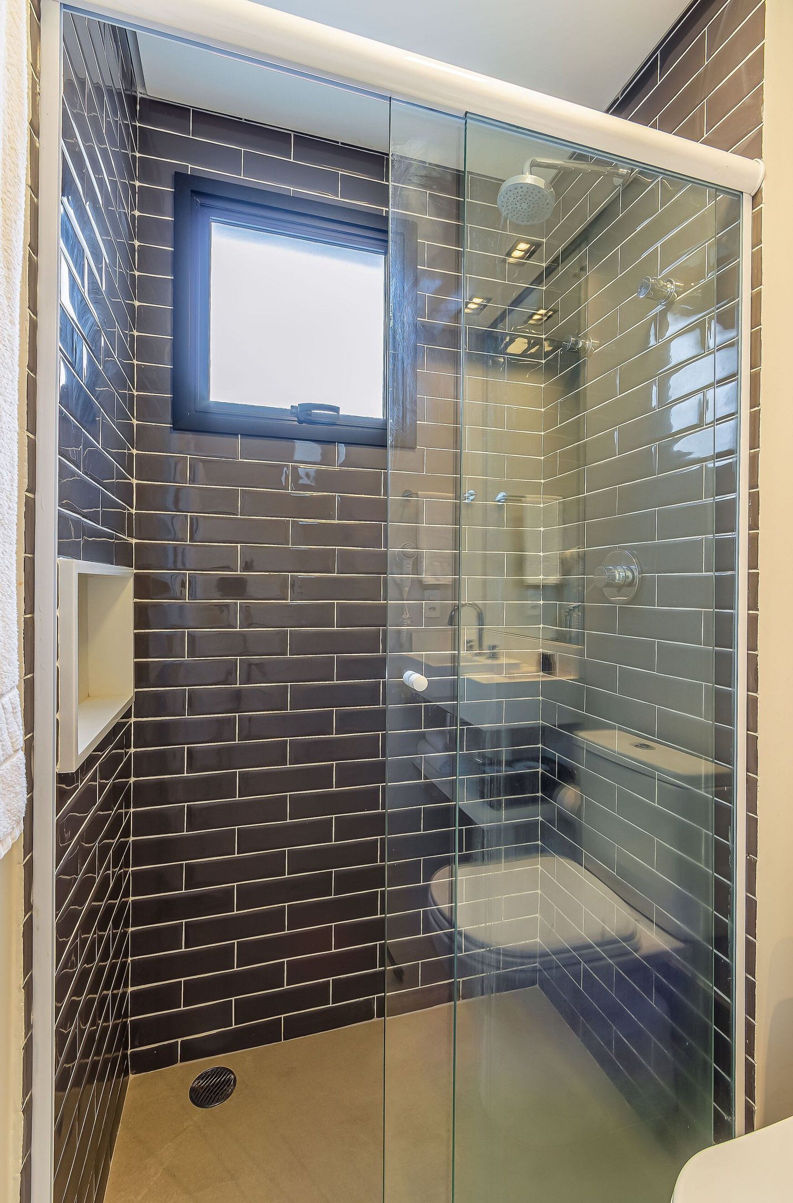 A modern bathroom with a glass shower enclosure, dark tiled walls, and a window above the sink.