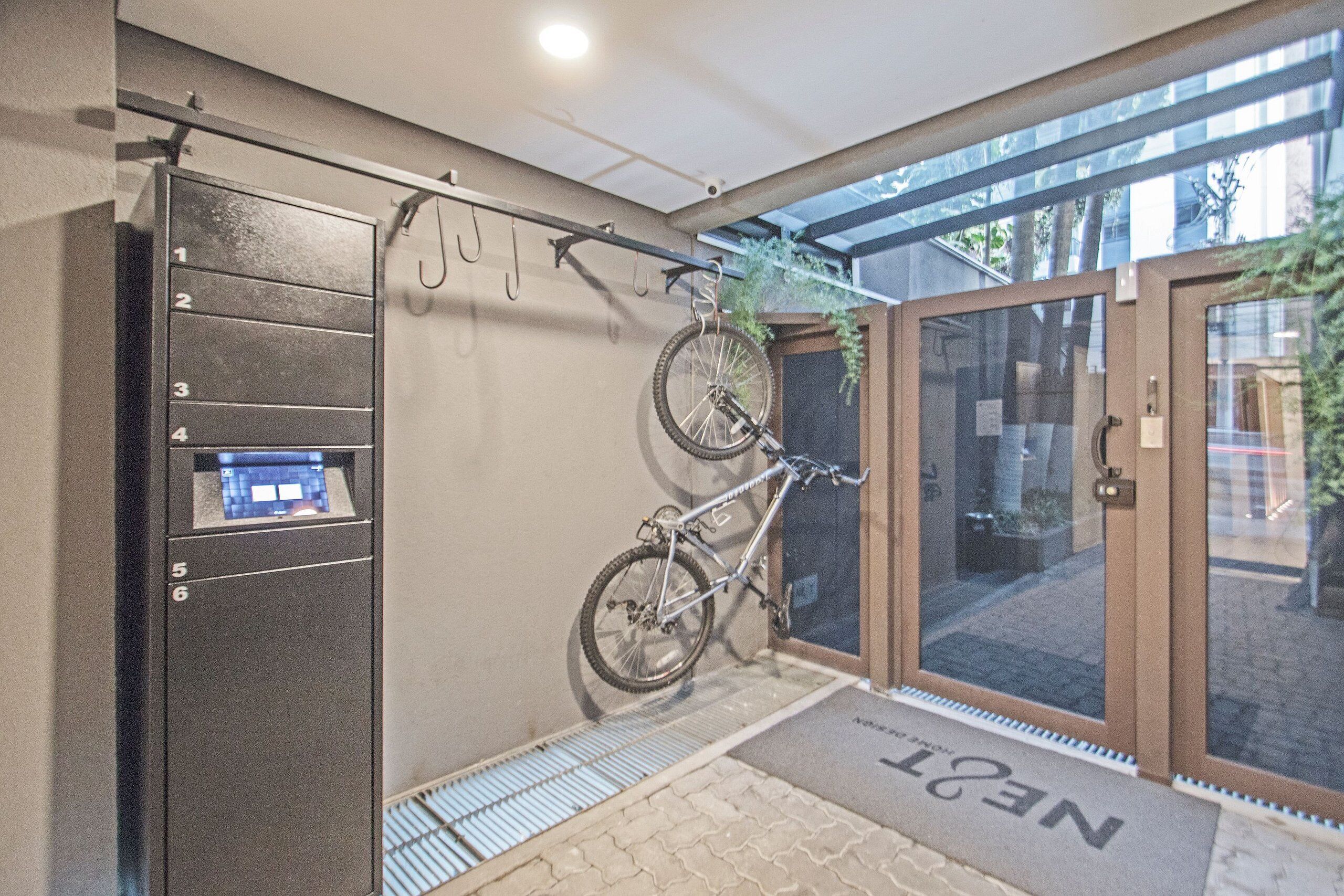 Entrada de un hotel con un aparca bicis, un panel de botones numerado y una puerta de cristal que da al exterior.