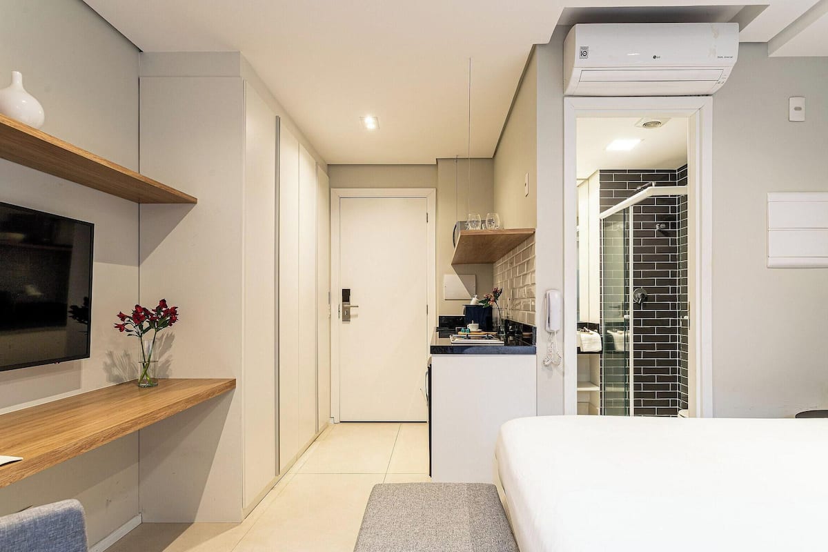A modern hotel room with a TV, a bedside table, a bed, a bathroom with a shower, and a kitchen area.