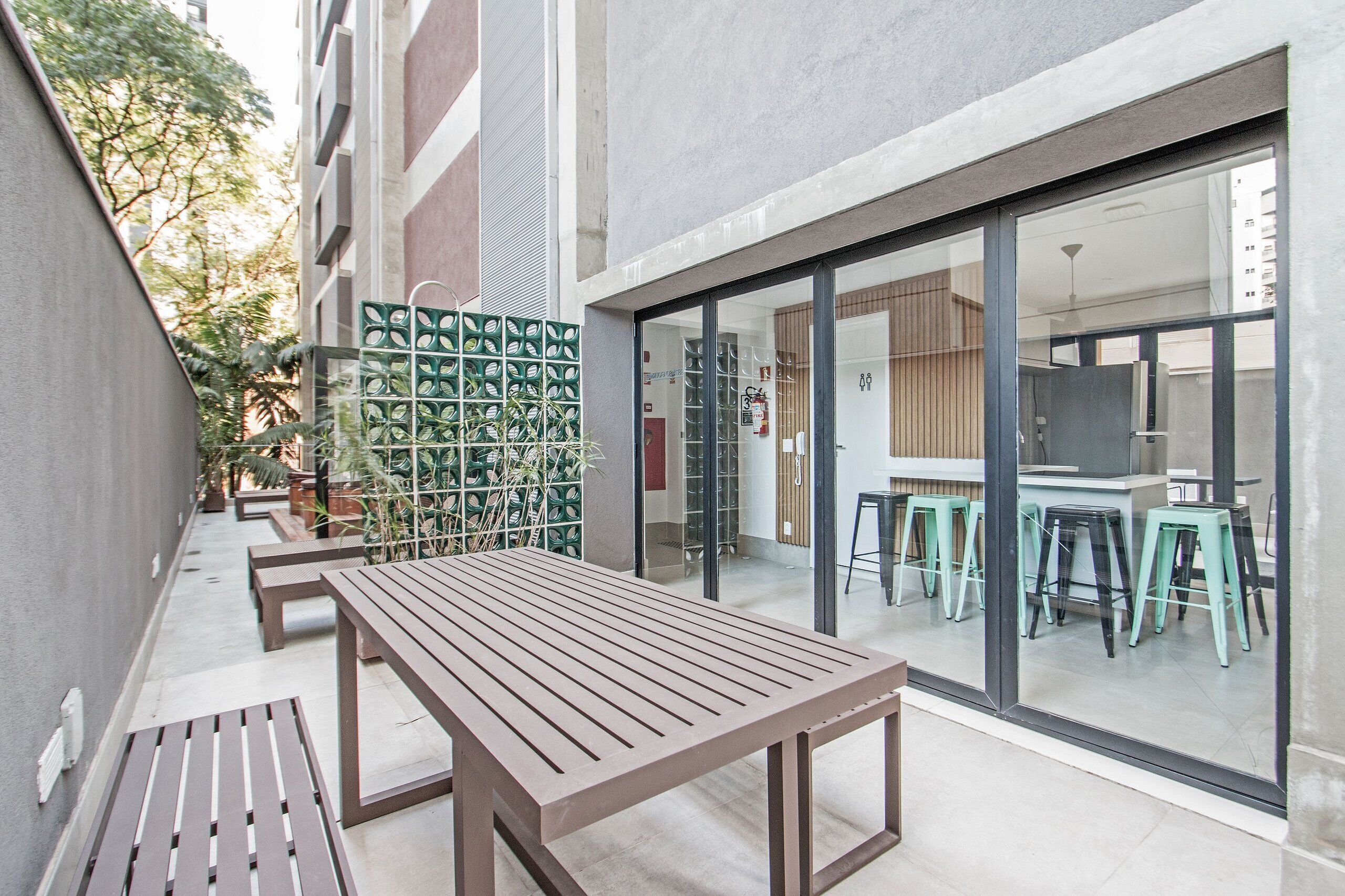 An outdoor patio with wooden benches, a green plant wall, and a glass door leading to an interior space with stools.