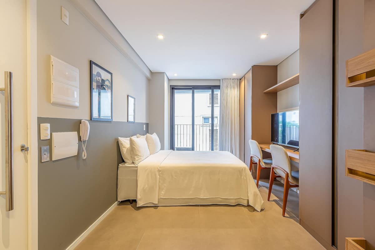 A hotel room with a bed, a desk with a chair, a TV, a window with curtains, and a door.