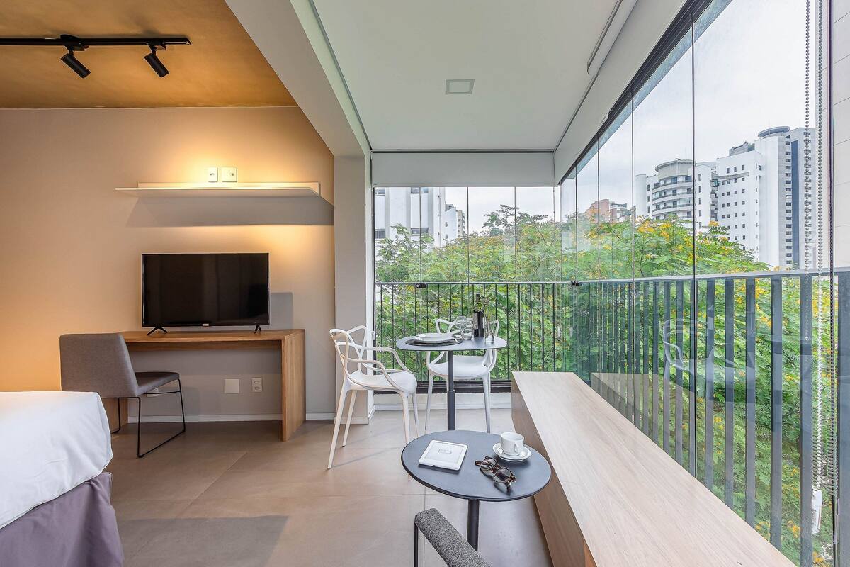 A modern room with a bed, a desk with a TV, a balcony with a table and chairs, and a city view.