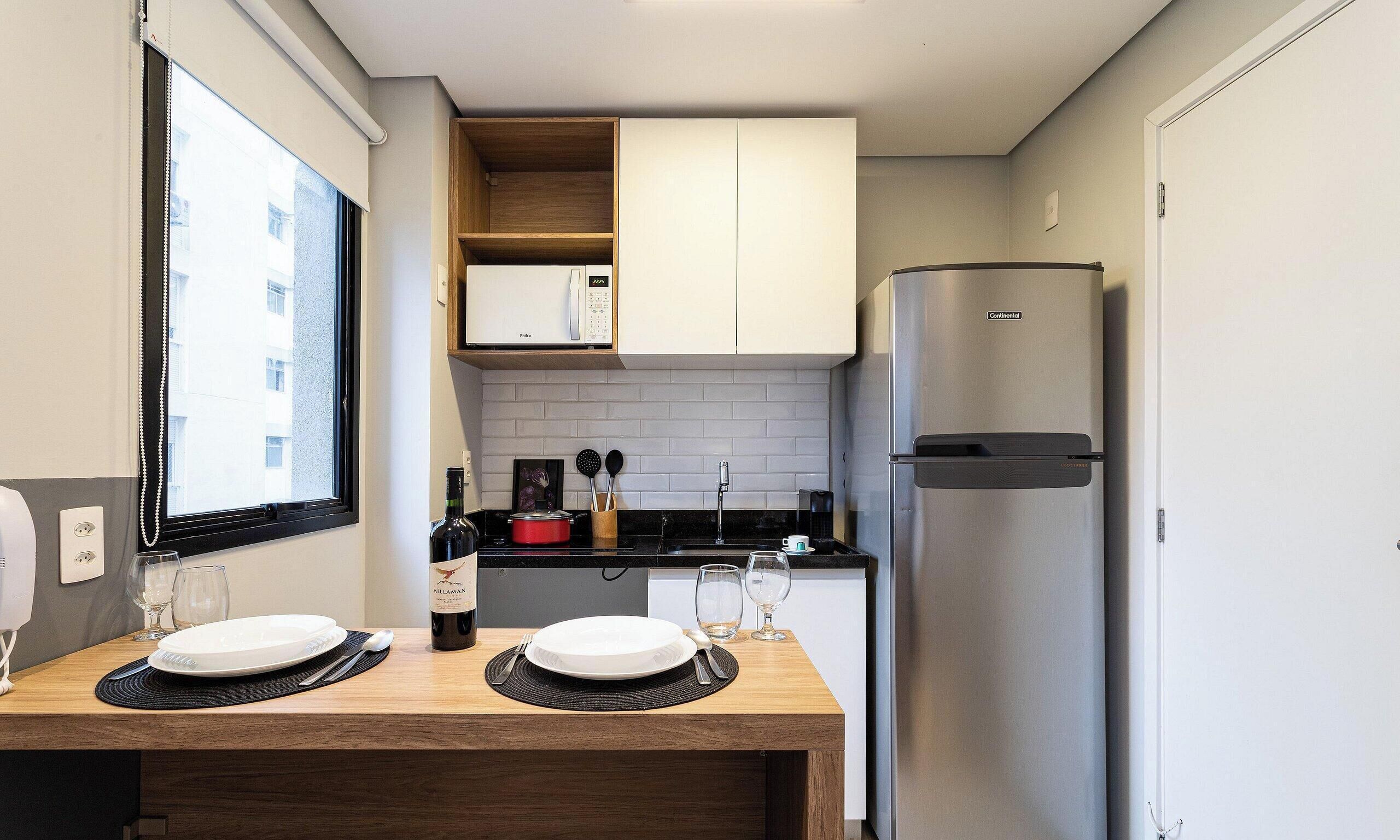 A compact kitchen with a built-in refrigerator, a window with blinds, and a dining area with two plates and wine glasses.
