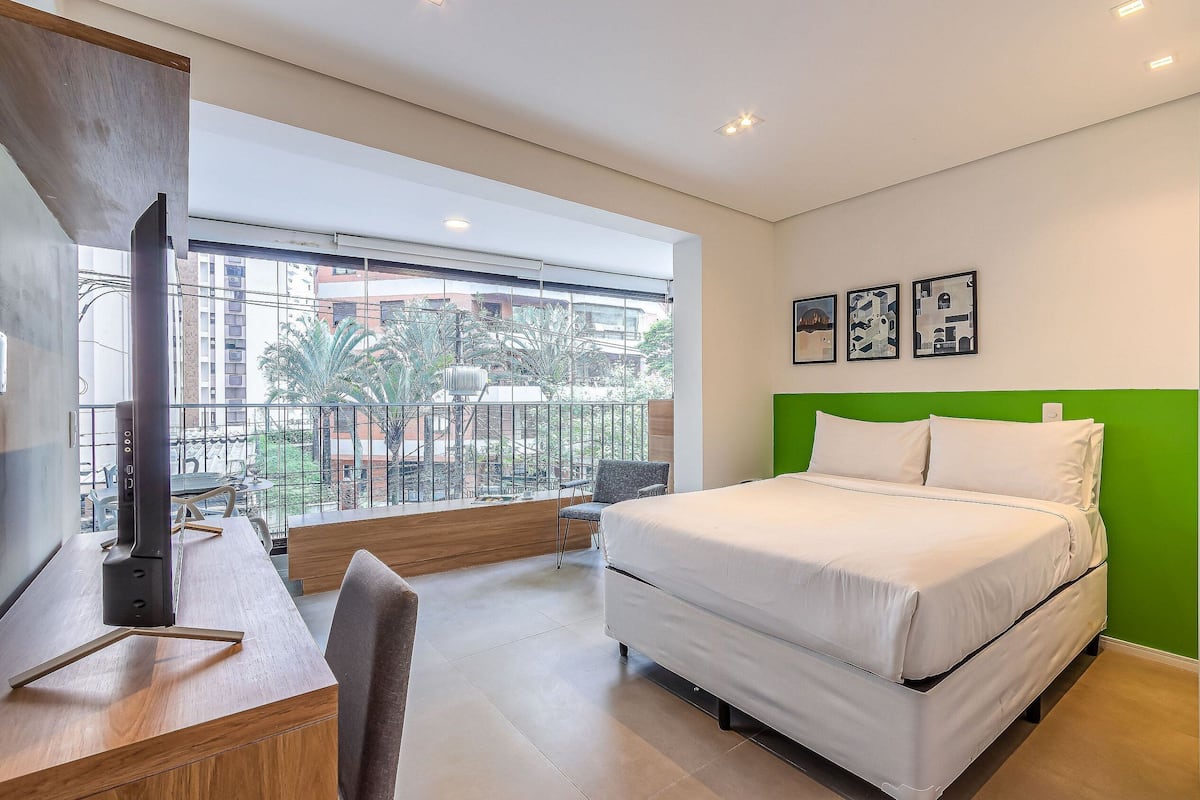 A hotel room with a bed, a desk with a computer, and a balcony with a view of buildings and trees.