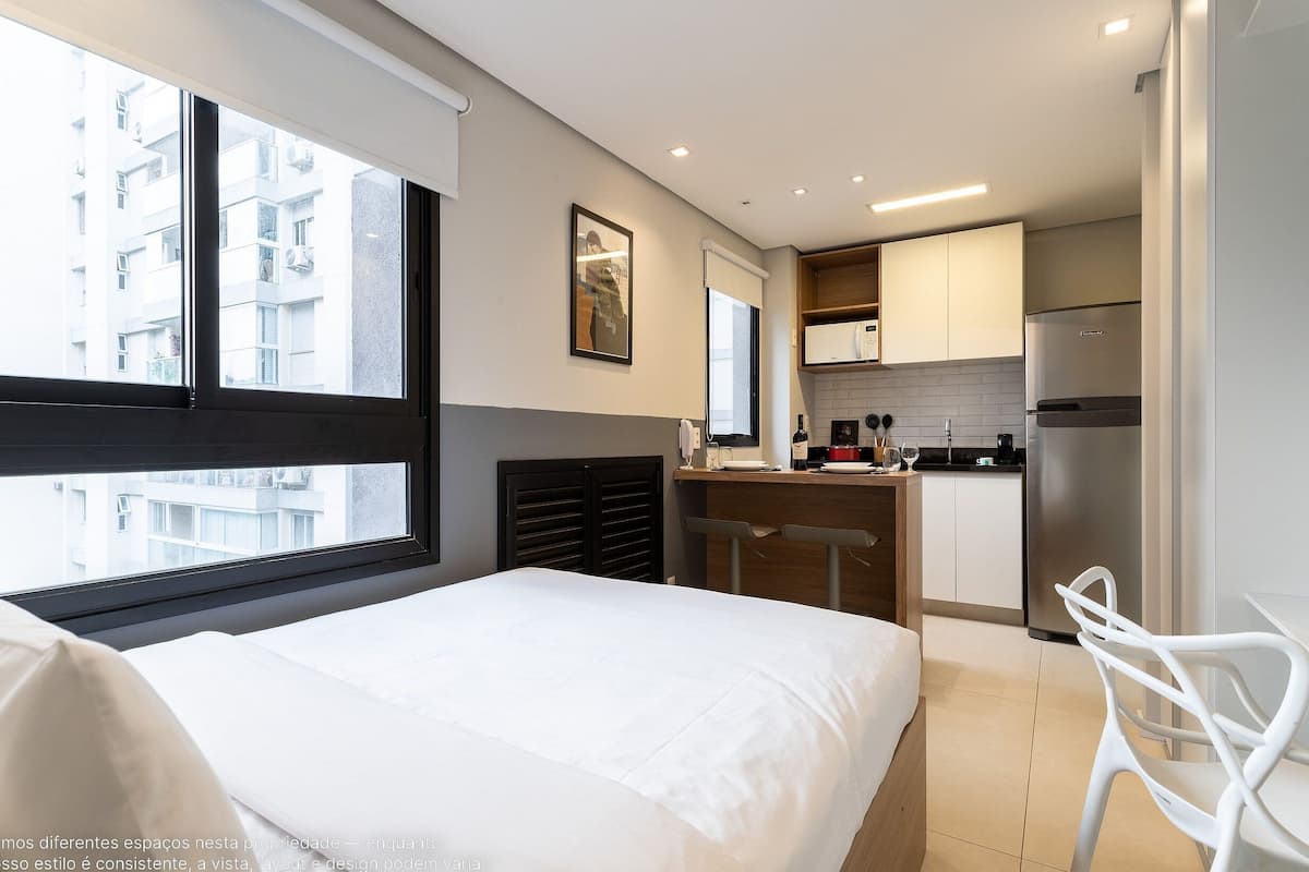 A bedroom with a large window, a bed, a desk, and a kitchen area with a refrigerator.