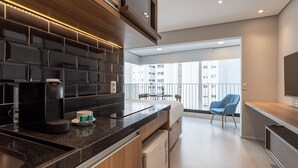 A modern kitchen with a black backsplash, a coffee maker, and a small refrigerator. There is a balcony with a chair and a table. - Next Home Design by Viva - Apartamentos em prédio próximo ao Parque do Ibirapuera (São Paulo)