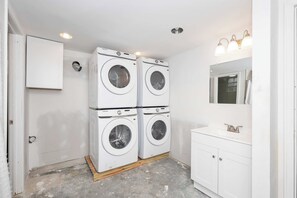 Une buanderie équipée de quatre machines à laver superposées, d’un meuble blanc, d’un évier avec un miroir et de spots encastrés au plafond.