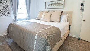 Une chambre à coucher bien rangée, avec un lit, des rideaux, un tableau encadré et une porte dotée d’un porte-manteau.
