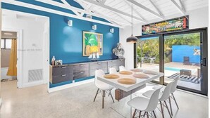 A modern kitchen with a blue accent wall, a dining area with white chairs, and a view of a pool through large glass doors.