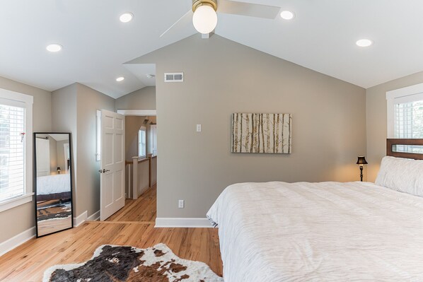 A bedroom with a large bed, a ceiling fan, recessed lighting, a mirror, and a door leading to another room.