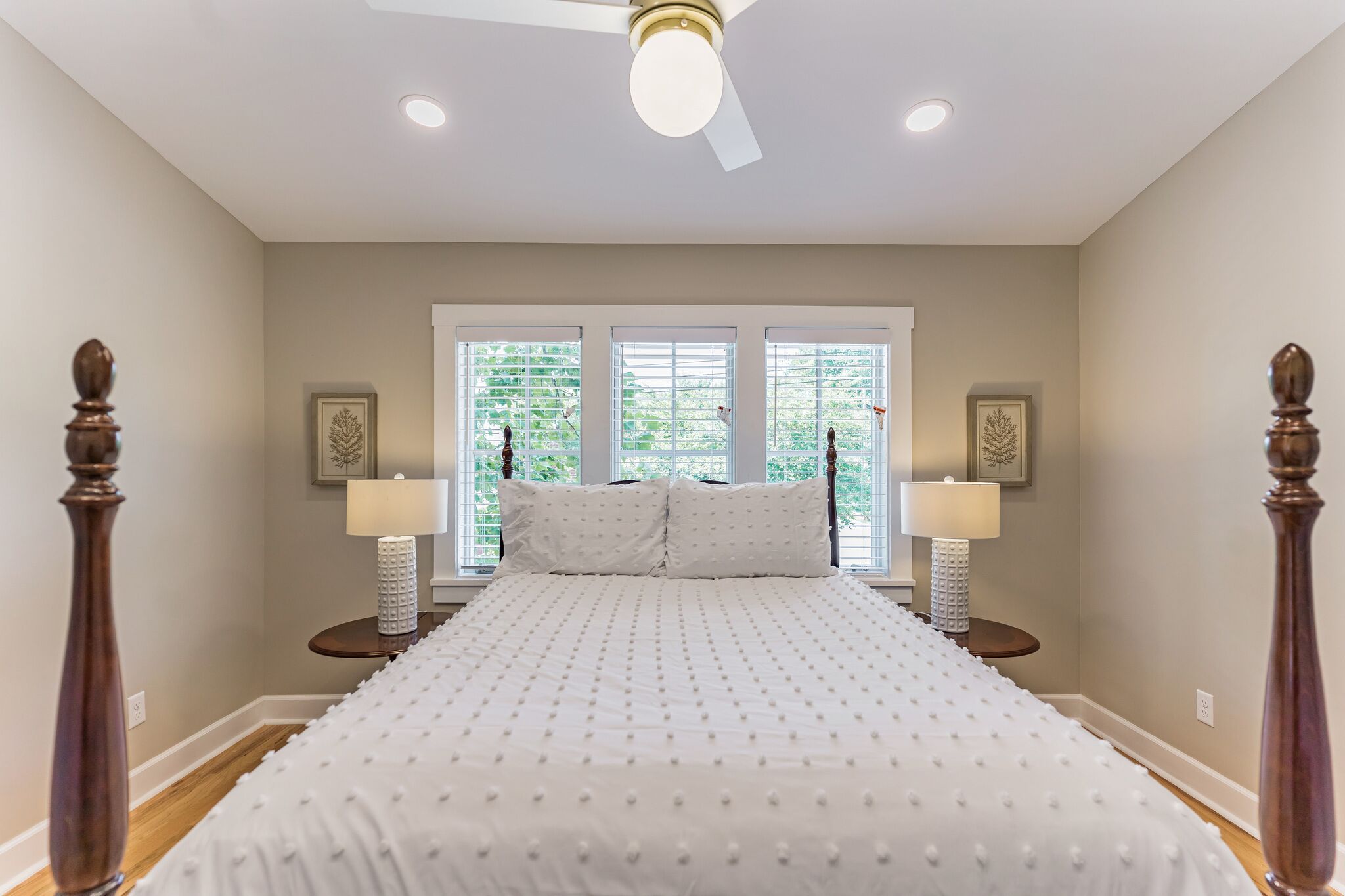 A bedroom with a large bed, two bedside tables with lamps, a ceiling fan, and windows with blinds.