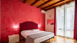 Ein Schlafzimmer mit roter Wand, Holzboden, einem Bett mit weißer Bettwäsche, einem Nachttisch und einem Fenster mit Vorhängen.