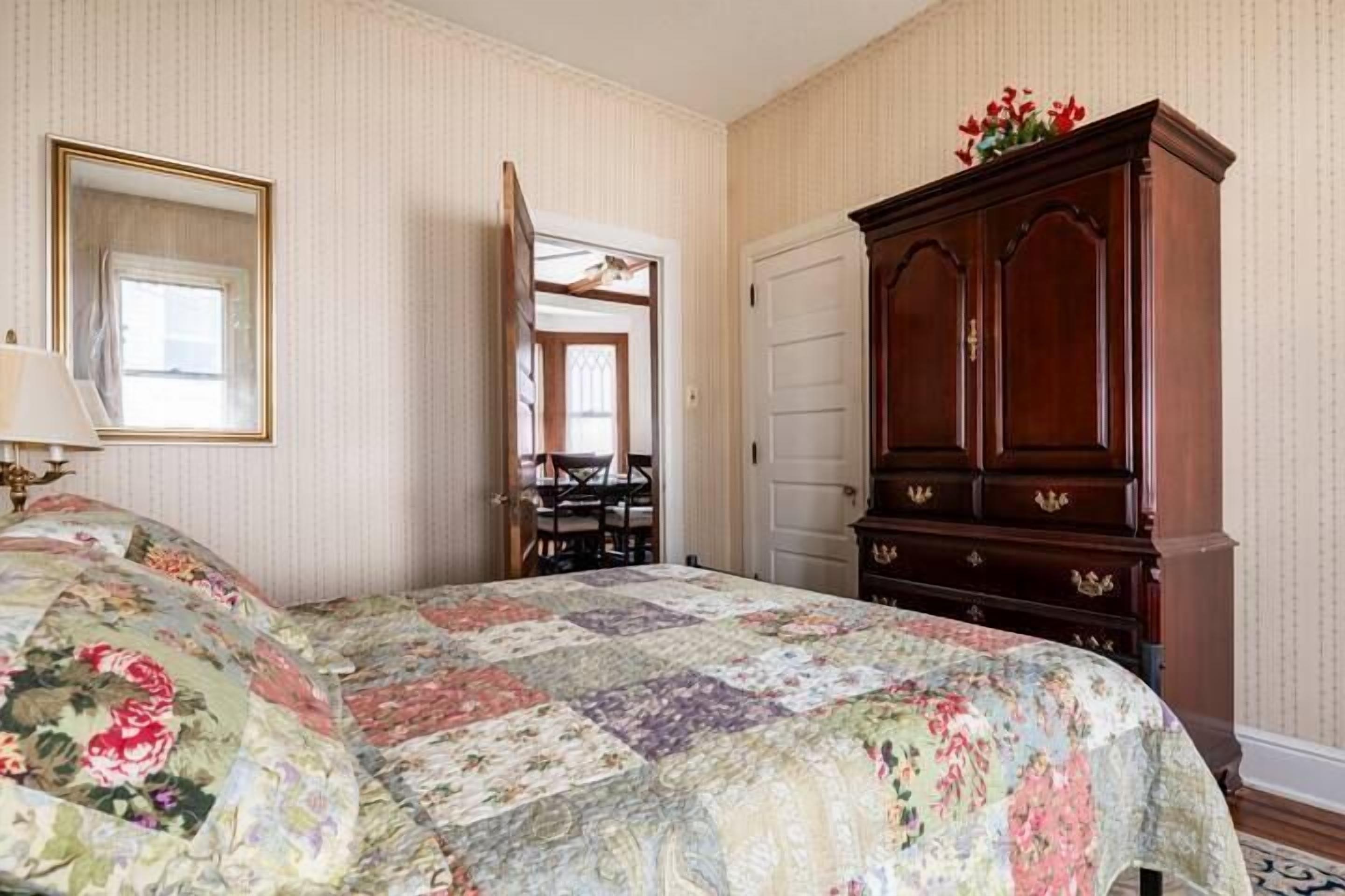 A bedroom with a floral quilt, a wooden dresser, a mirror, and a door leading to another room.