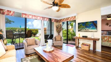 A spacious living room with a wooden coffee table, comfortable seating, a ceiling fan, and large windows offering a view of the outdoors.