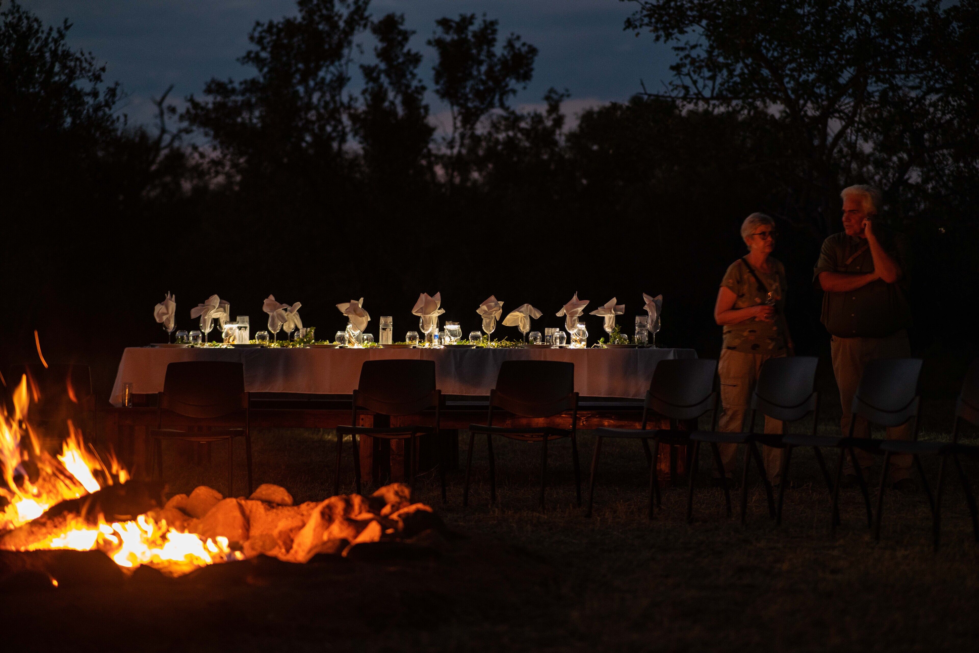 An outdoor dining area with a long table set for a meal, surrounded by chairs, and a fire pit in the foreground.