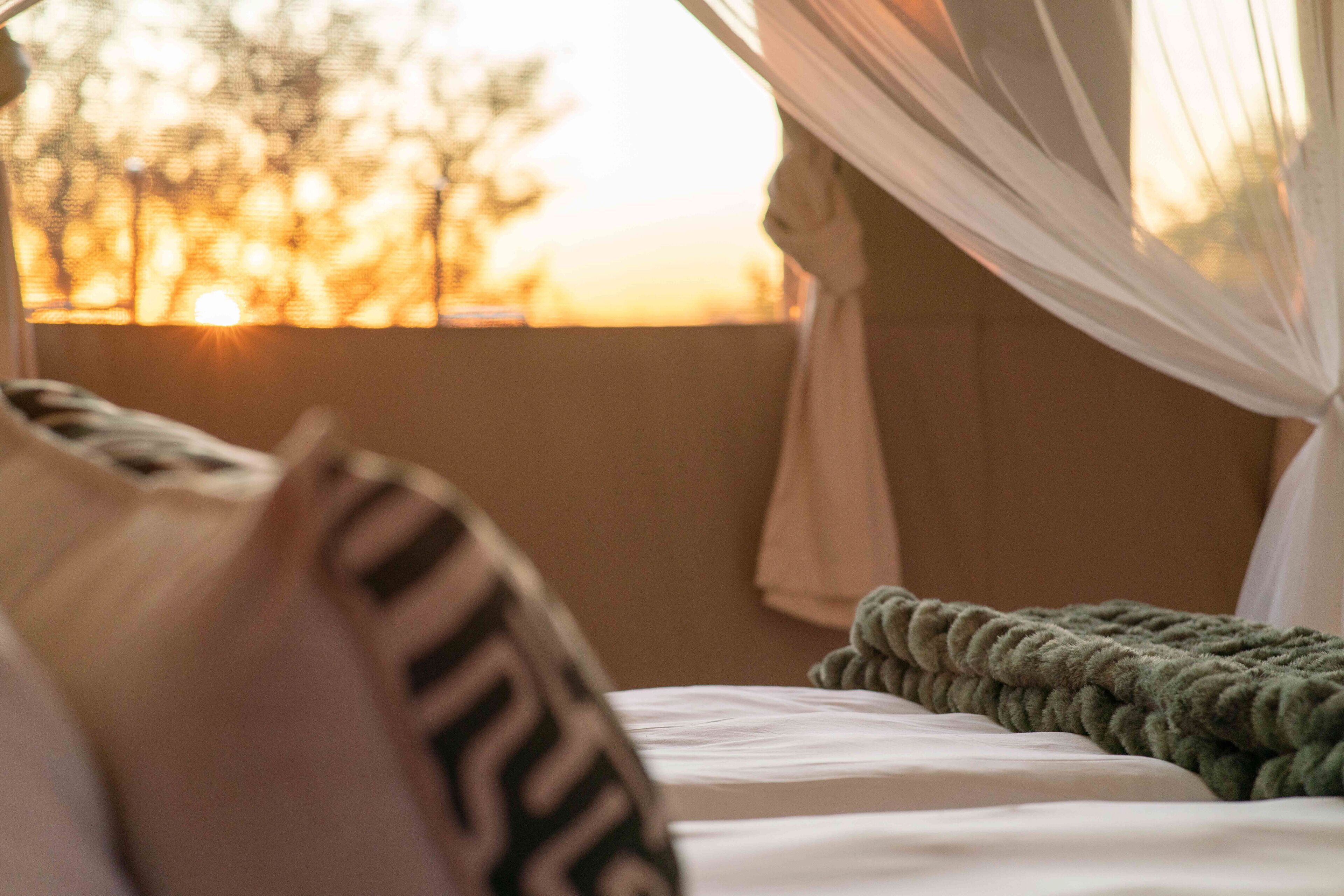 Una cama con una manta verde y un almohadón blanco con texto negro, una ventana con cortinas de encaje y vistas al atardecer.