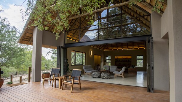 A spacious outdoor deck with wooden flooring and seating, leading to a modern interior with large windows and a view of the surrounding landscape. - Cape Vulture Lodge (Hoedspruit)