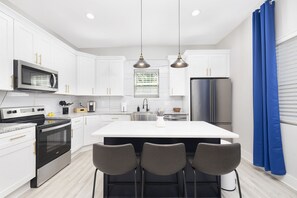 A modern kitchen with white cabinets, stainless steel appliances, a central island with bar stools, pendant lighting, and a window with blue curtains. - Downtown Modern Living! (Concord)