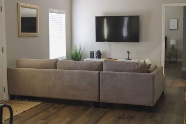 A living room with a beige sectional sofa, a flat-screen TV mounted on the wall, a small side table with a lamp, and a potted plant. - Cozy & Private home w/ in-ground pool (Warner Robins)