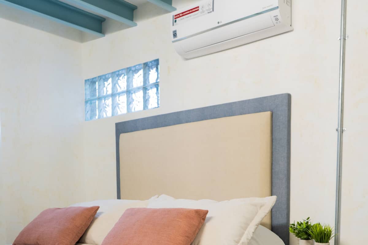 A bedroom with a bed, pillows, a bedside table with a plant, a wall-mounted air conditioner, and a window with blinds.