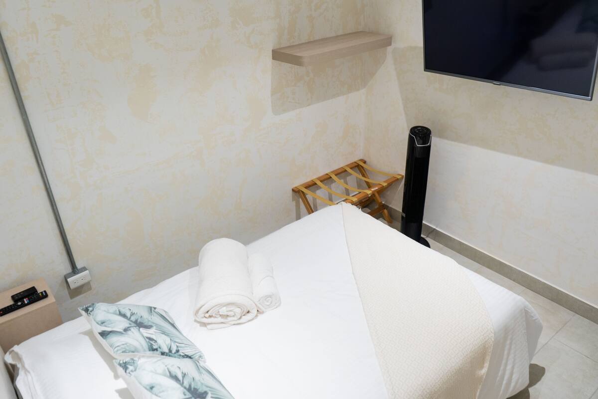 A neatly made bed with a rolled towel, a wooden chair, a black vase, and a mounted TV.