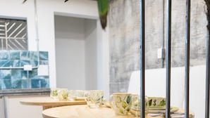 Un área de comedor moderna con mesas y sillas de madera, una planta en maceta y vista a la cocina al fondo.