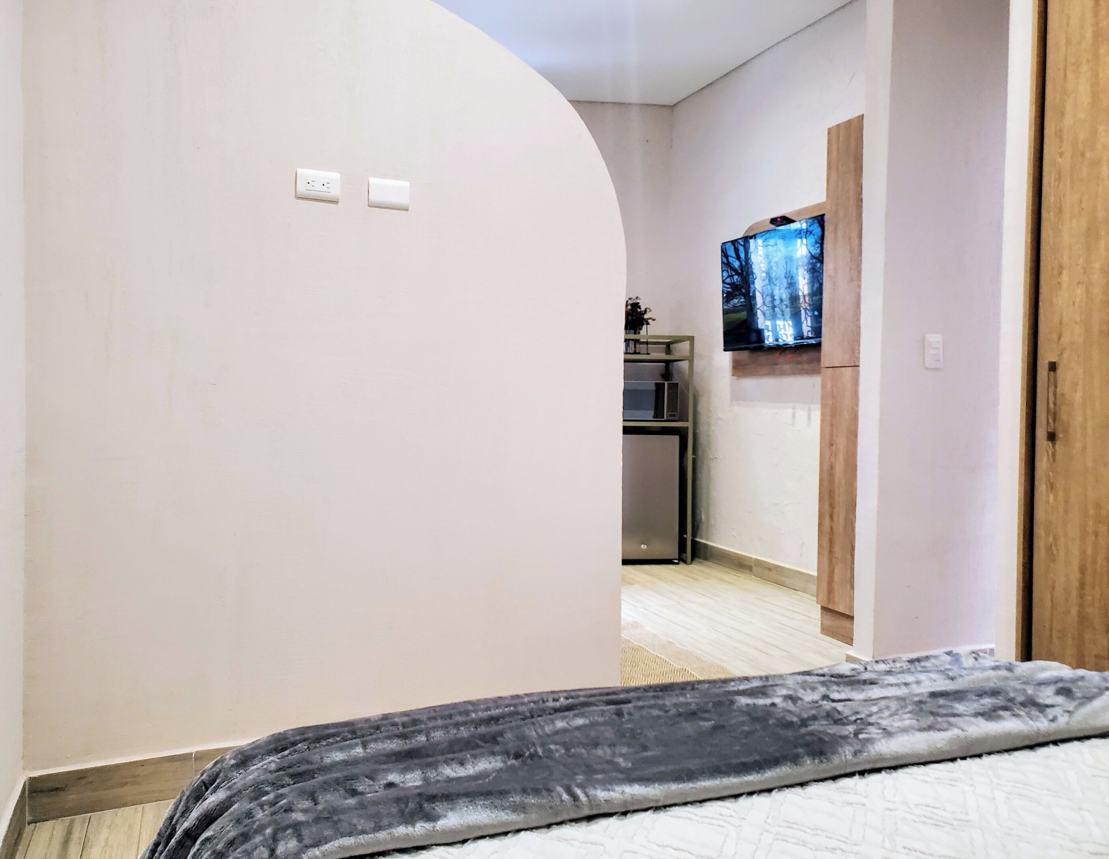 A bedroom with a bed, a TV mounted on the wall, and a kitchenette in the background.