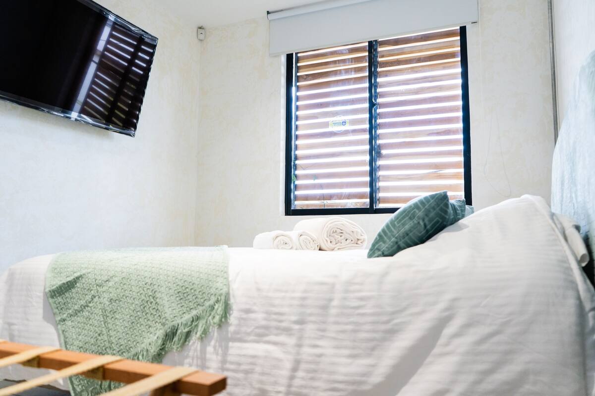 A bedroom with a bed, a window with blinds, a TV mounted on the wall, and a green throw blanket.