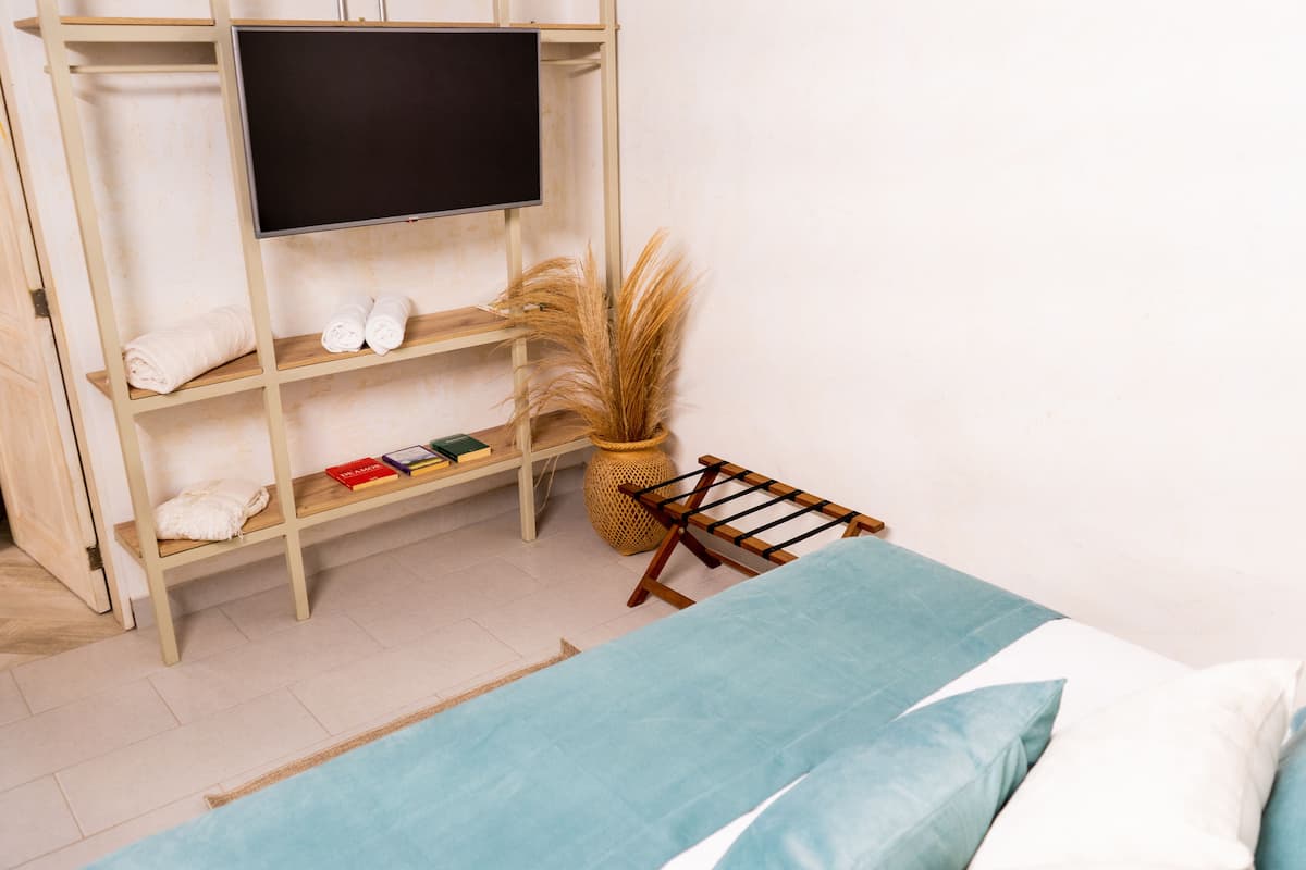 A room with a bed, a TV mounted on a wooden shelf, a small table with books, and a woven basket.